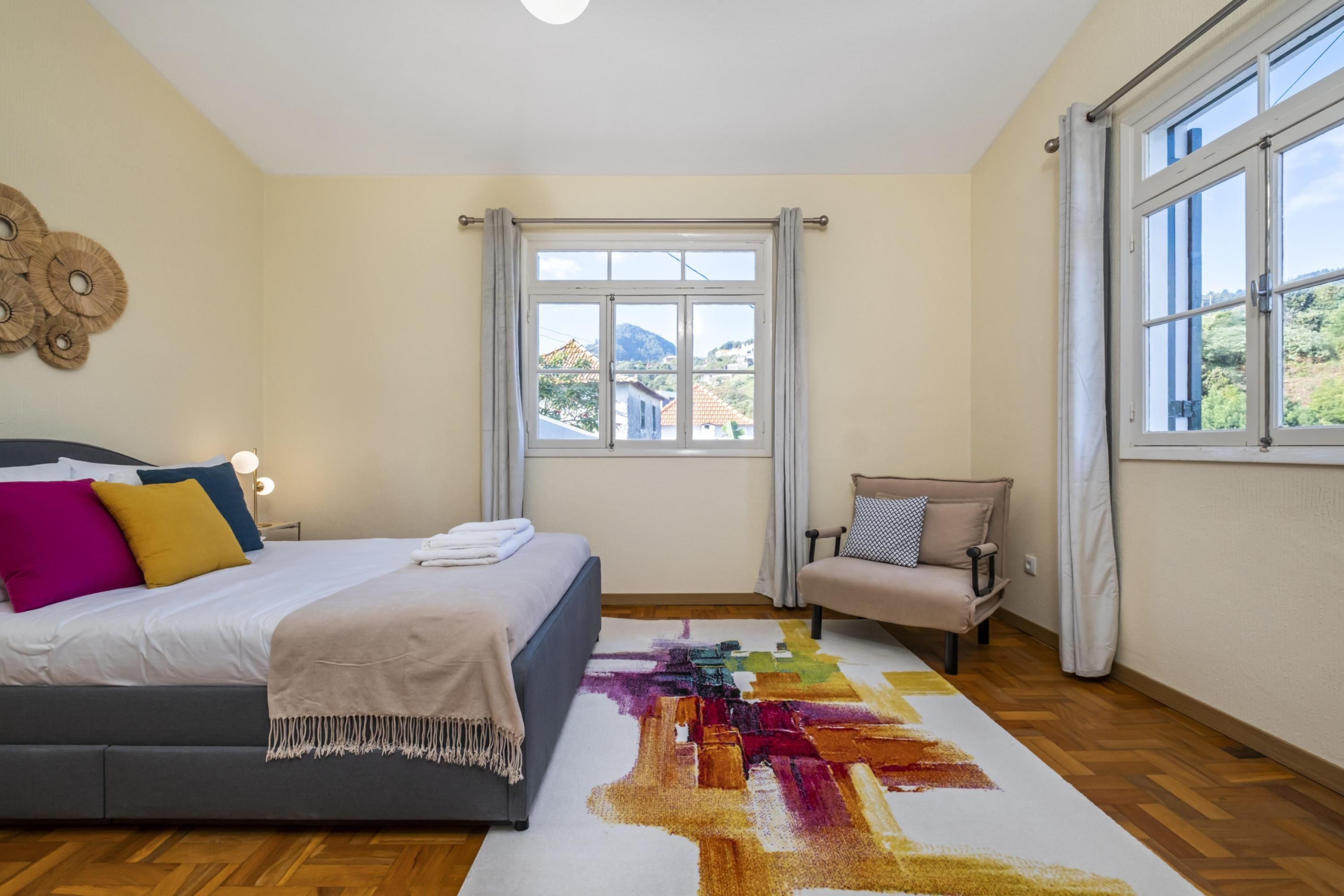A bedroom with a bed, a chair, a window with curtains, and a view of buildings outside.