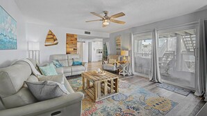 Ein geräumiges Wohnzimmer mit Deckenventilator, großem Fenster mit Vorhängen, Couchtisch aus Holz und einem gemusterten Teppich.