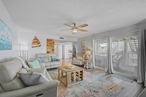 A spacious living room with a ceiling fan, a large window with curtains, a wooden coffee table, and a patterned rug.