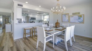 A modern kitchen with white cabinetry, a central island, and a dining area with white wicker chairs and a glass table. - Luxury Gulf-Front Condo w/ Balcony Views | 3BR/2BA | Pool | Beach Access | Sleeps 6 | Great Sunsets | Anna Maria Island (Holmes Beach)