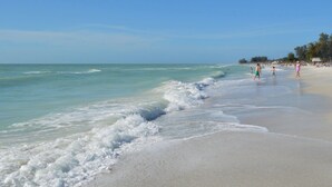 A beach with clear turquoise water, gentle waves, and people walking along the shore. - Luxury Gulf-Front Condo w/ Balcony Views | 3BR/2BA | Pool | Beach Access | Sleeps 6 | Great Sunsets | Anna Maria Island (Holmes Beach)