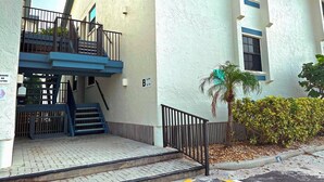 A residential building with a blue staircase leading to a gated entrance, a palm tree, and a hedge. - Luxury Gulf-Front Condo w/ Balcony Views | 3BR/2BA | Pool | Beach Access | Sleeps 6 | Great Sunsets | Anna Maria Island (Holmes Beach)