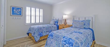 Un dormitorio con dos camas, una ventana con persianas, un cuadro colgado en la pared y una mesita de noche con lámpara.