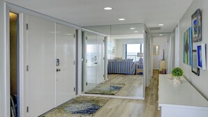 A hallway with a mirrored closet door, a bed with striped bedding, and a small plant on a table. - Luxury Gulf-Front Condo w/ Balcony Views | 3BR/2BA | Pool | Beach Access | Sleeps 6 | Great Sunsets | Anna Maria Island (Holmes Beach)