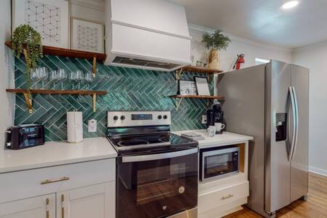 A modern kitchen with a stainless steel refrigerator, a double oven, and a microwave. There are wooden shelves above the counter and a green patterned backsplash.
