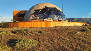A geodesic dome structure with a wooden exterior and a large glass facade, situated on a grassy hill with a clear sky.