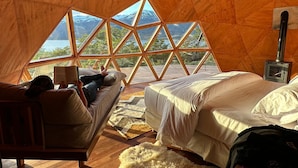 A cozy room with a wooden ceiling, a bed, a sofa, and a large window offering a mountain view.