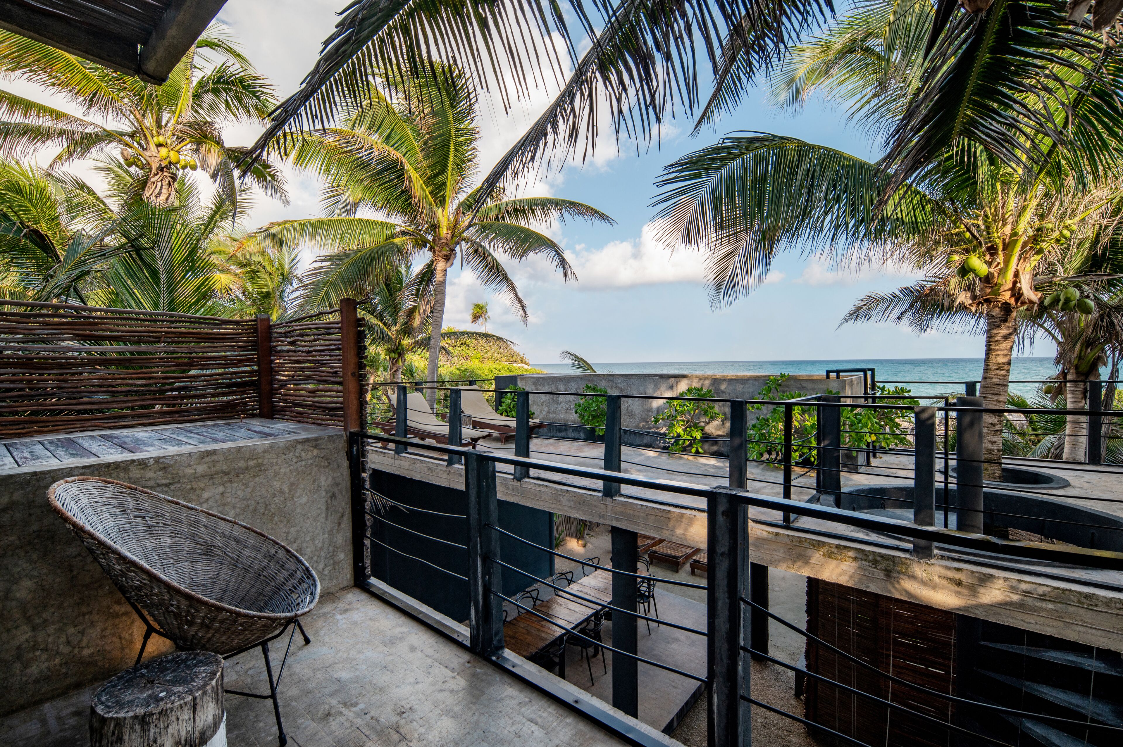 A balcony with wicker chairs, a concrete floor, and a view of the ocean and palm trees.