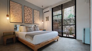 A bedroom with a wooden bed, a nightstand, a bedside table, and a large window with a view of a garden. - Tropicalito Tulum (Tulum)