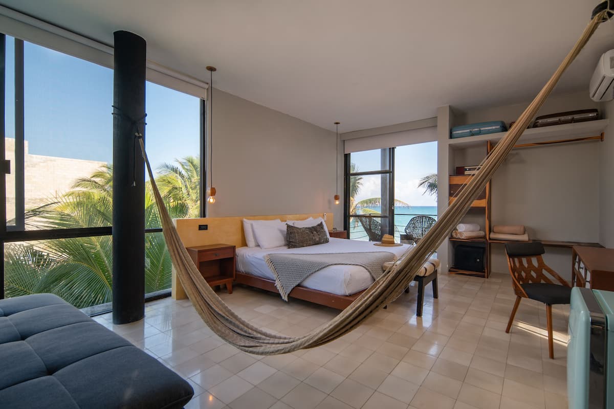 Un dormitorio moderno con una cama grande, una hamaca, un escritorio y vistas a palmeras a través de amplias ventanas.