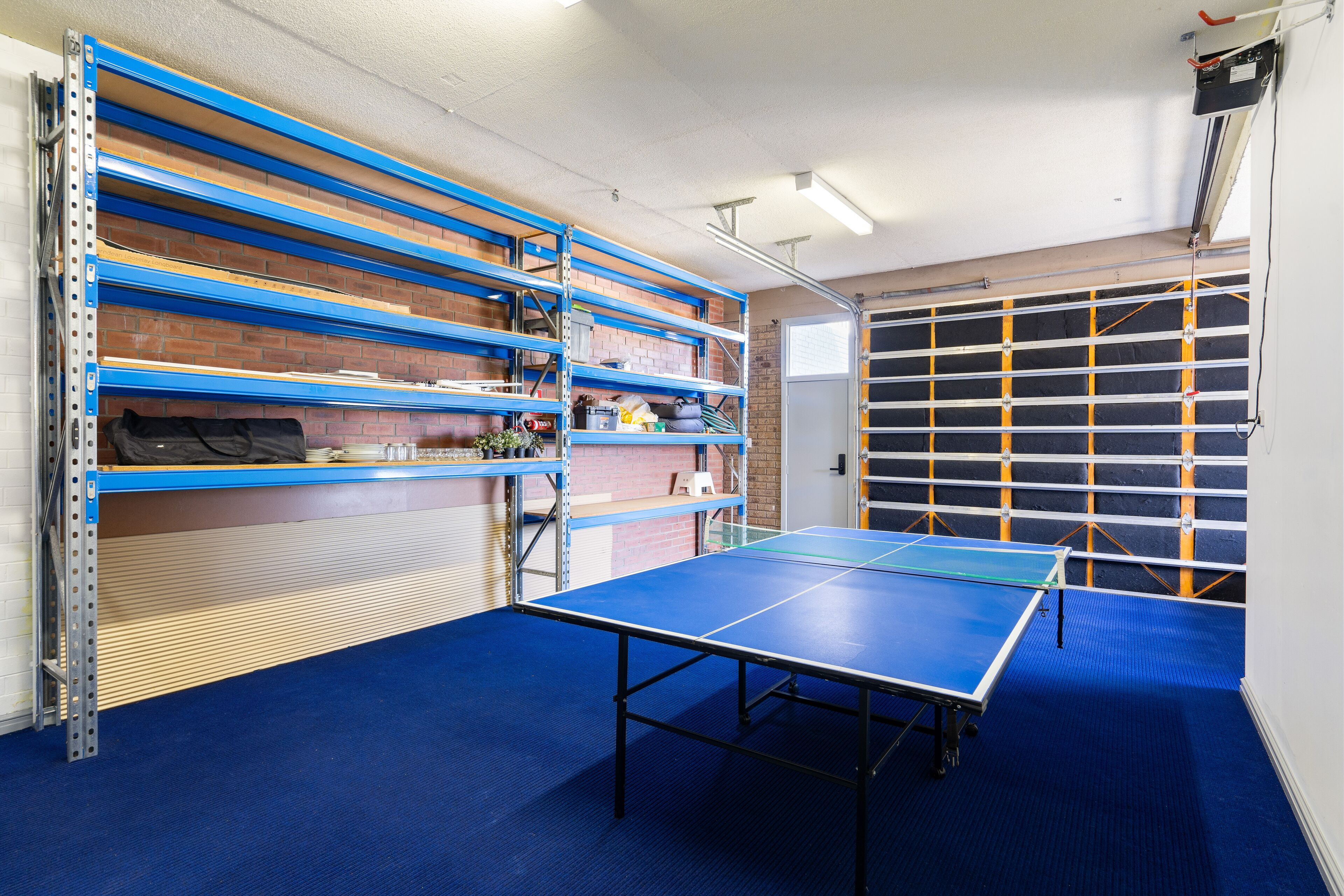 A room with a ping pong table, shelving units, and a garage door.