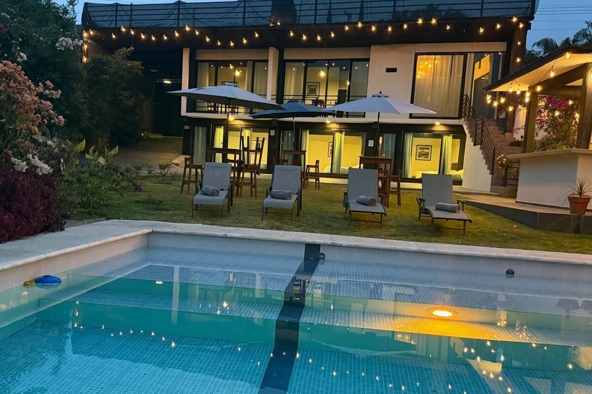 Une piscine entourée de chaises longues et de parasols, elle-même bordée d’une pelouse bien entretenue et d’une maison illuminée de guirlandes lumineuses.