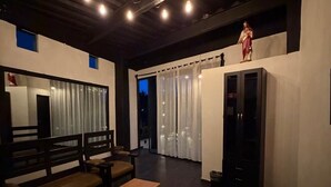 Ein modernes Wohnzimmer mit einer dunklen Holzbank, einem kleinen Holztisch und einem schwarzen Schrank. Ein Fenster mit Vorhängen und eine Statue auf dem Schrank sind ebenfalls zu sehen.