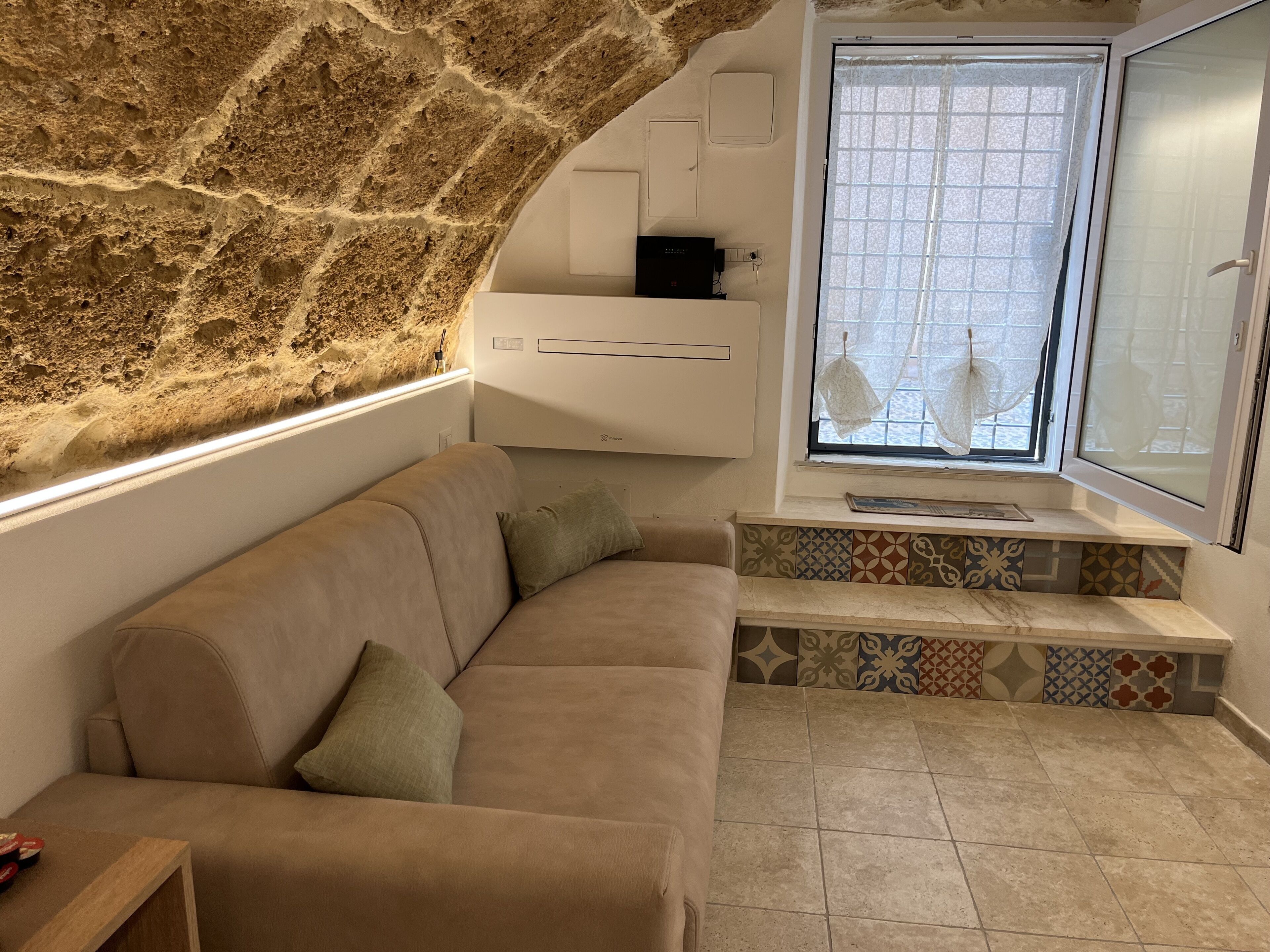 Zona d'estar acollidora amb una paret de pedra, un sofà beix, una finestra amb rajoles decorades i una taula petita amb un llibre.