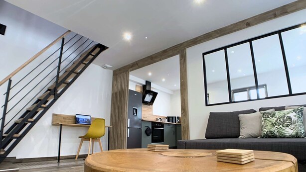 Ein modernes Wohnzimmer mit einem runden Holztisch, einer Couch mit Kissen, einem gelben Sessel, einer Küchenzeile und einer Treppe.