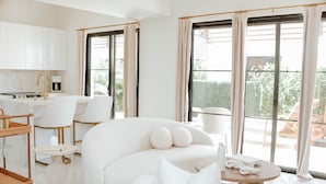 Een moderne, witte woonkamer met een bank, een klein rond tafeltje en een keukenhoek met witte kastjes en een koffieapparaat.