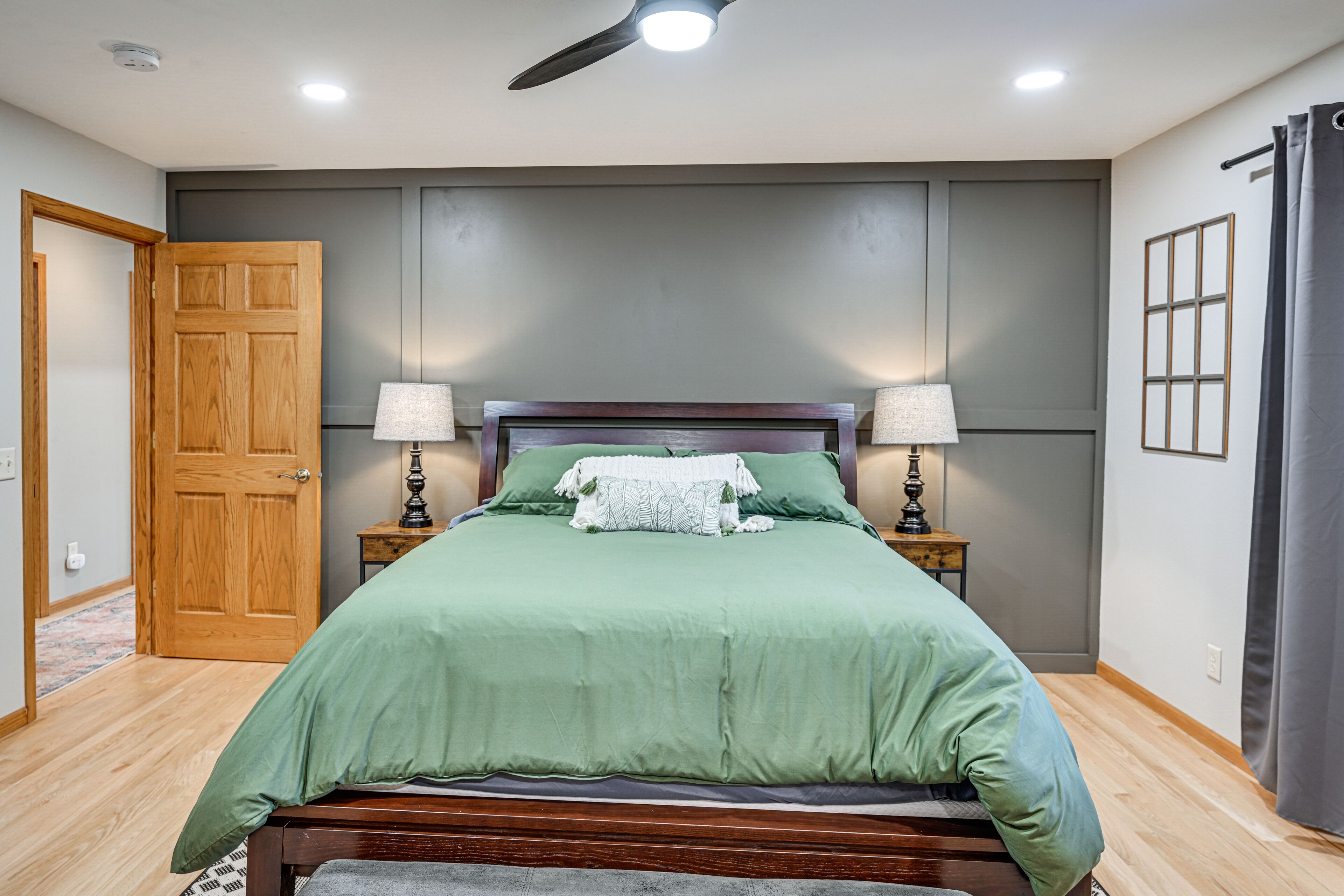 Une chambre à coucher avec un lit en bois, une literie verte, deux lampes de chevet, un ventilateur de plafond et une porte en bois.
