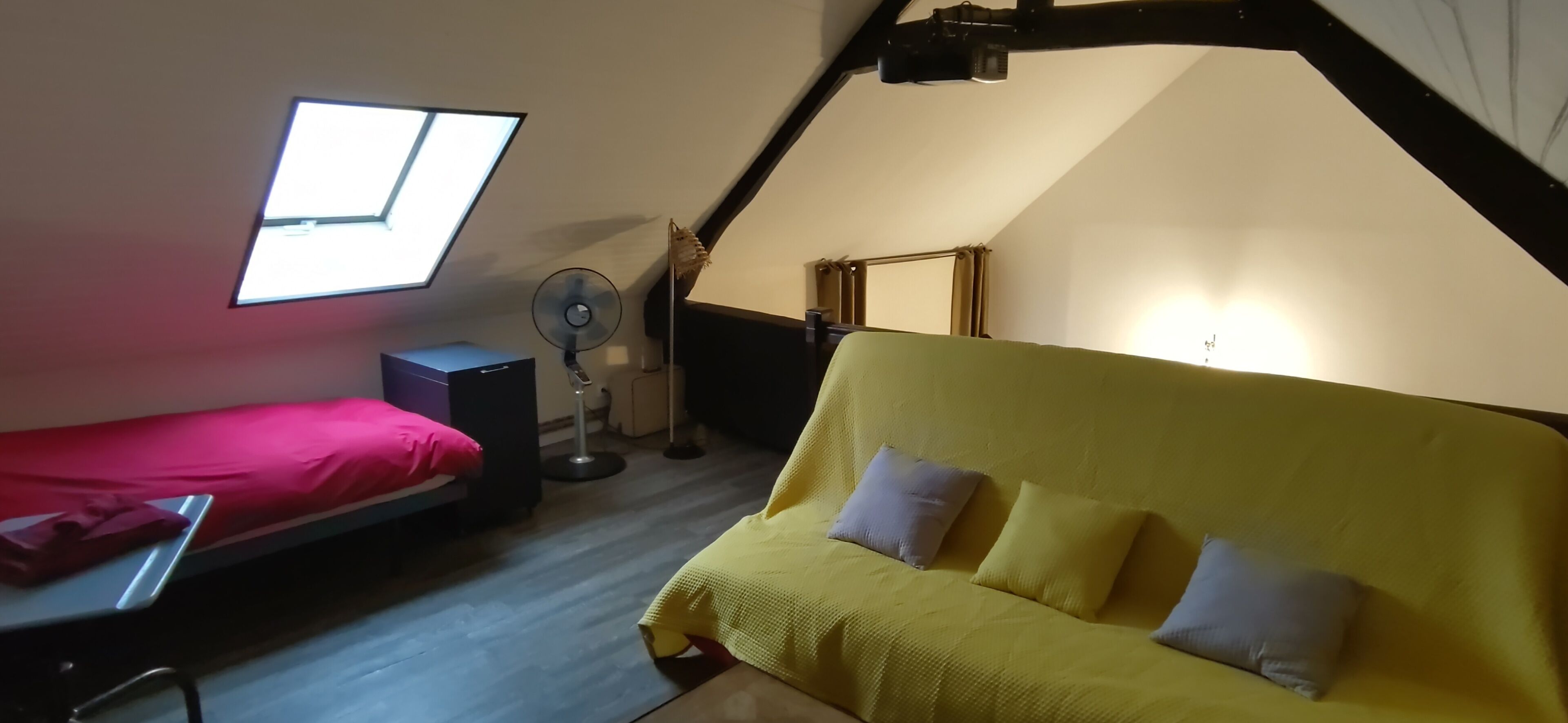 A small, cozy room with a sloped ceiling, a bed with a red mattress, a yellow sofa with cushions, a standing fan, and a window providing natural light.
