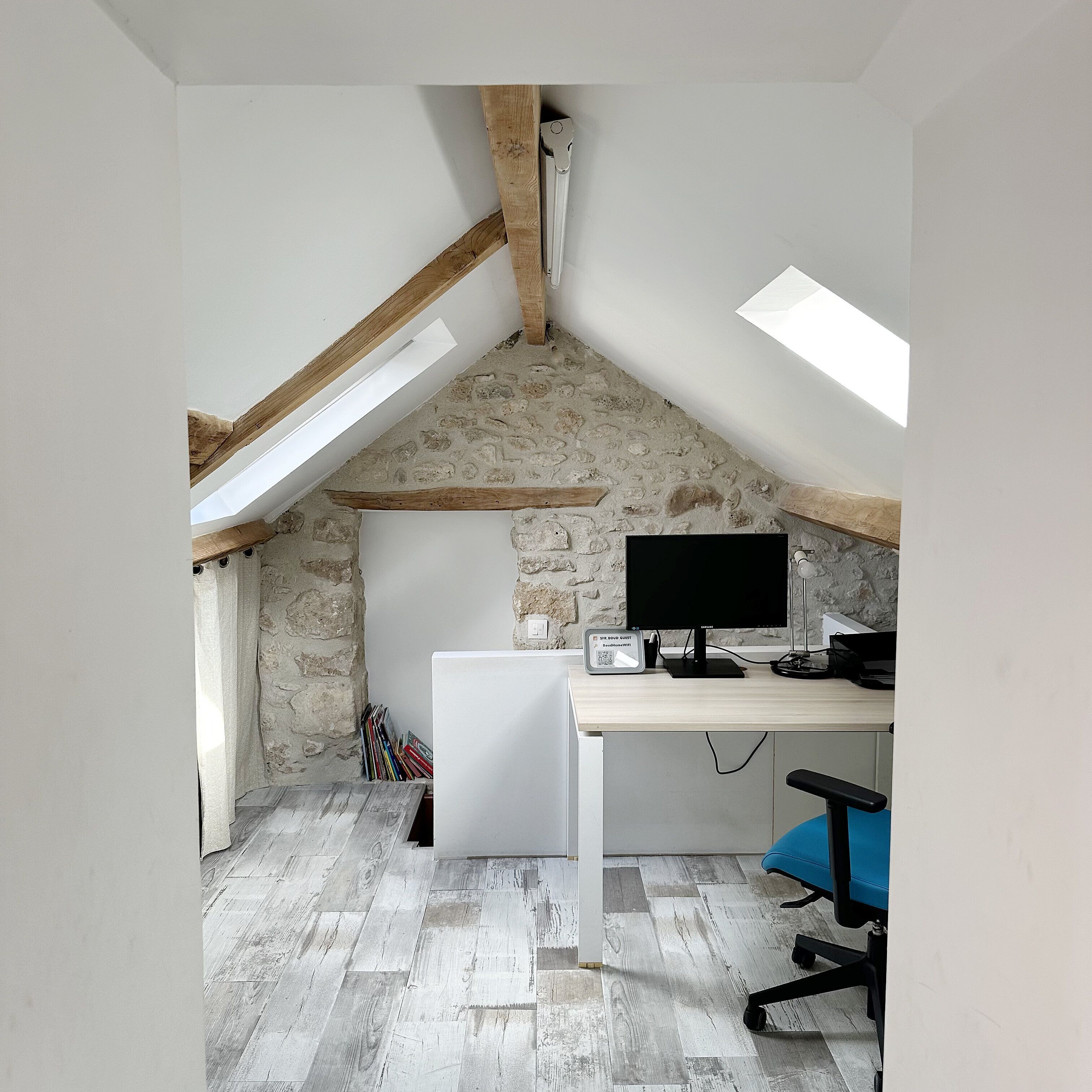 Una habitación con techo inclinado, un escritorio con ordenador, una silla y una pared de piedra.