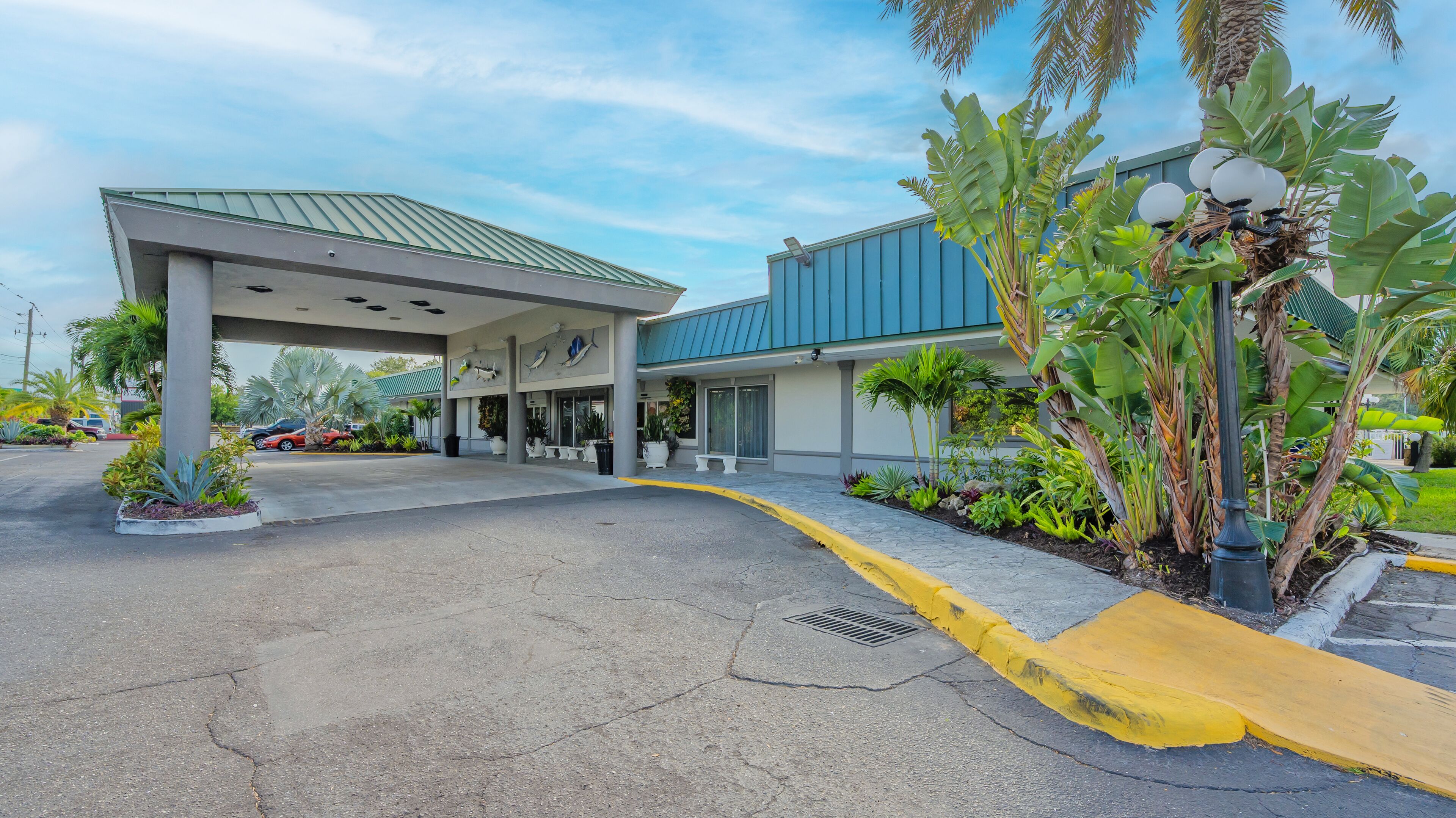 Entrada de hotel con estacionamiento techado, un edificio azul y jardines tropicales.