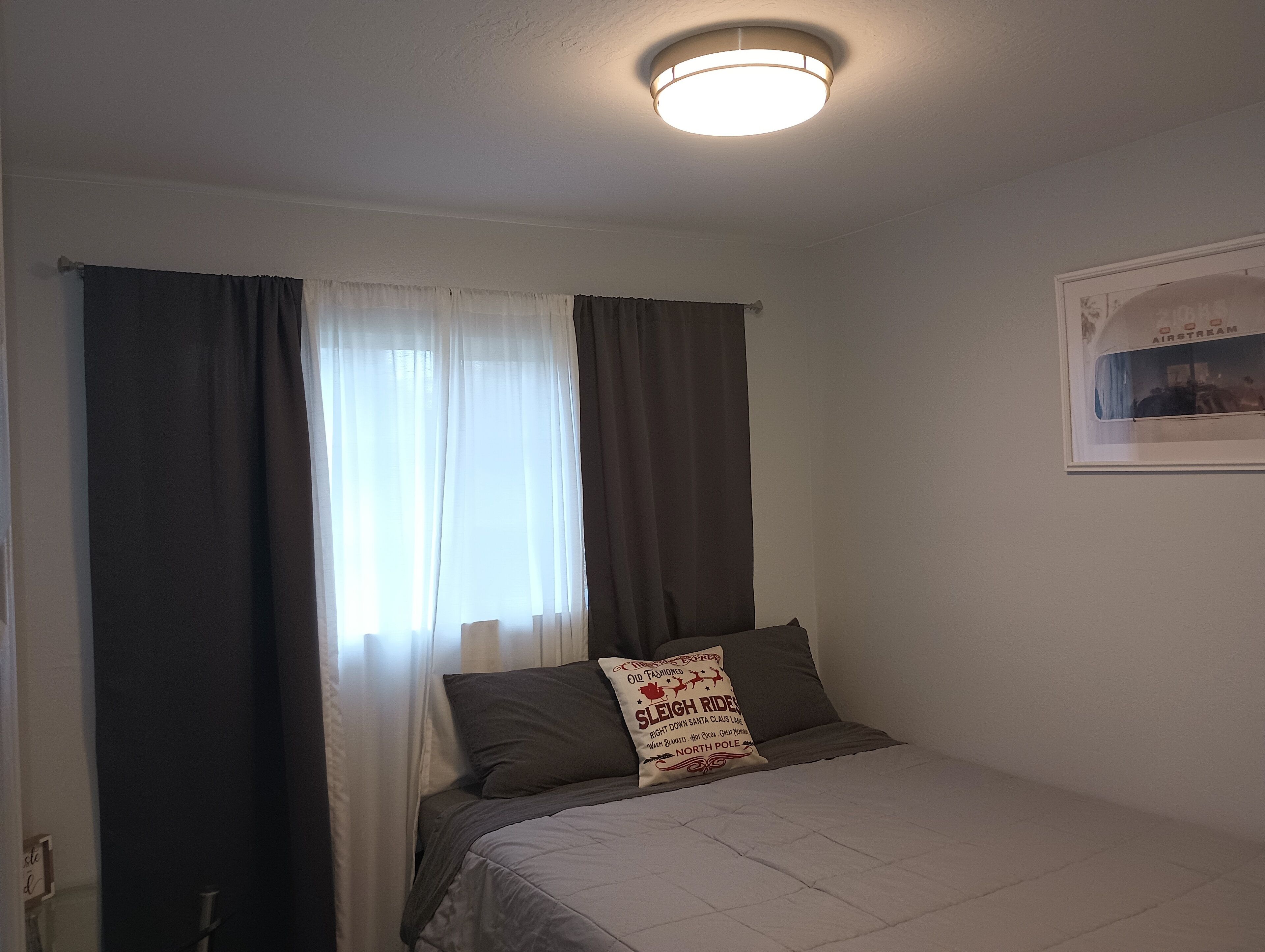 Um quarto com cama, cortinas, uma luminária no teto e um quadro na parede.