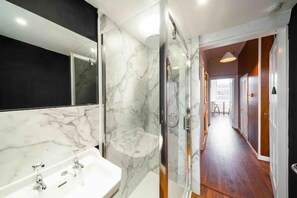 Een moderne badkamer met een marmeren wastafel, een douchecabine van glas en een houten gang die naar een andere kamer leidt.