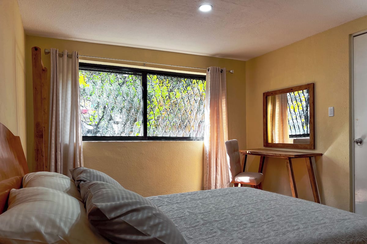 A bedroom with a bed, a chair, a small table, and a window with curtains.