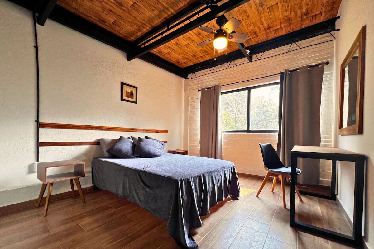 A bedroom with a wooden ceiling, a bed with a grey bedspread, a small bedside table, a chair, a desk, and a window with curtains.