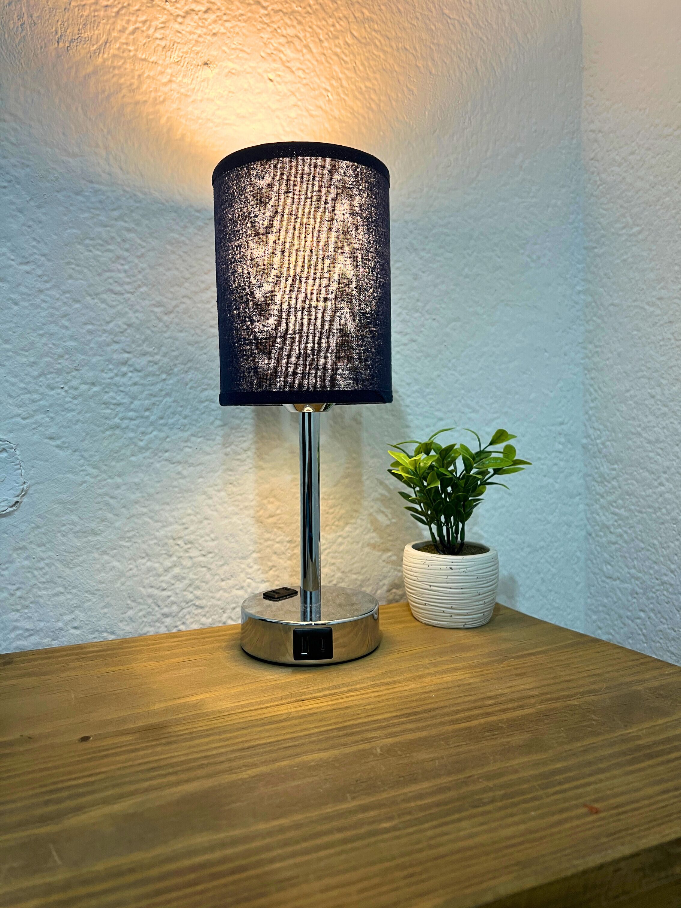 A lamp with a textured shade on a wooden surface, next to a potted plant.