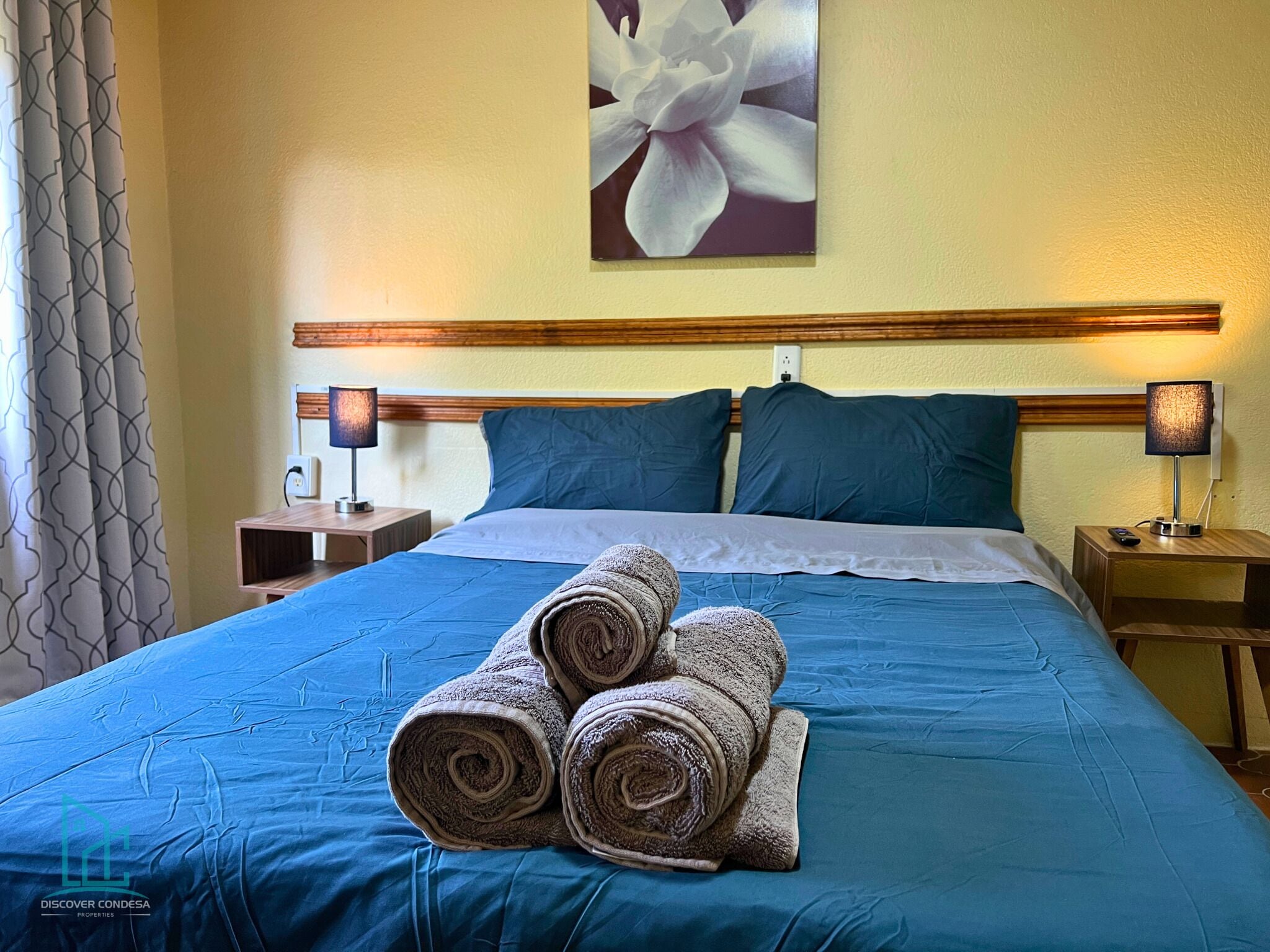 Une chambre d’hôtel avec un lit, deux tables de chevet, une lampe et un tableau représentant des fleurs accroché au mur.