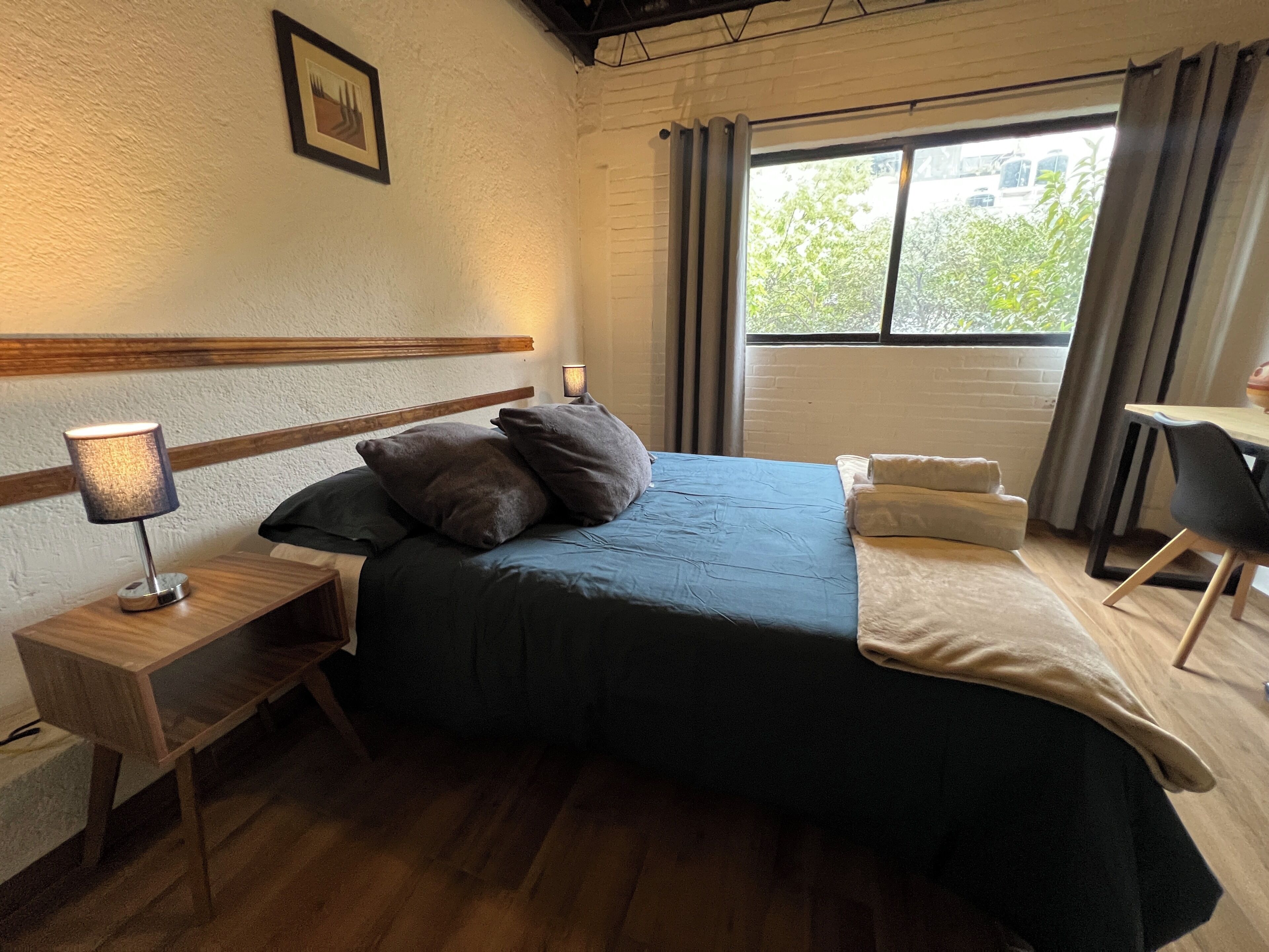 Habitación de hotel con cama, lámpara de noche, escritorio y ventana con cortinas.
