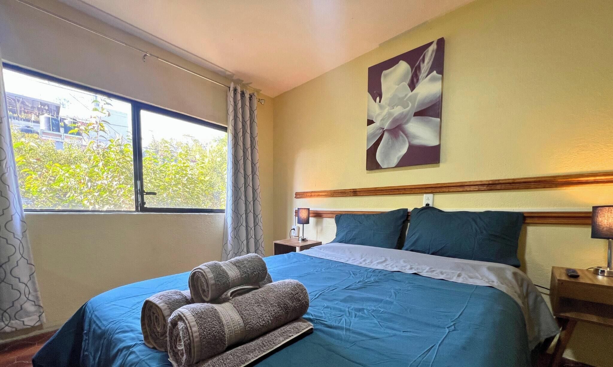 A hotel room with a bed, a window with curtains, a painting on the wall, and a nightstand.
