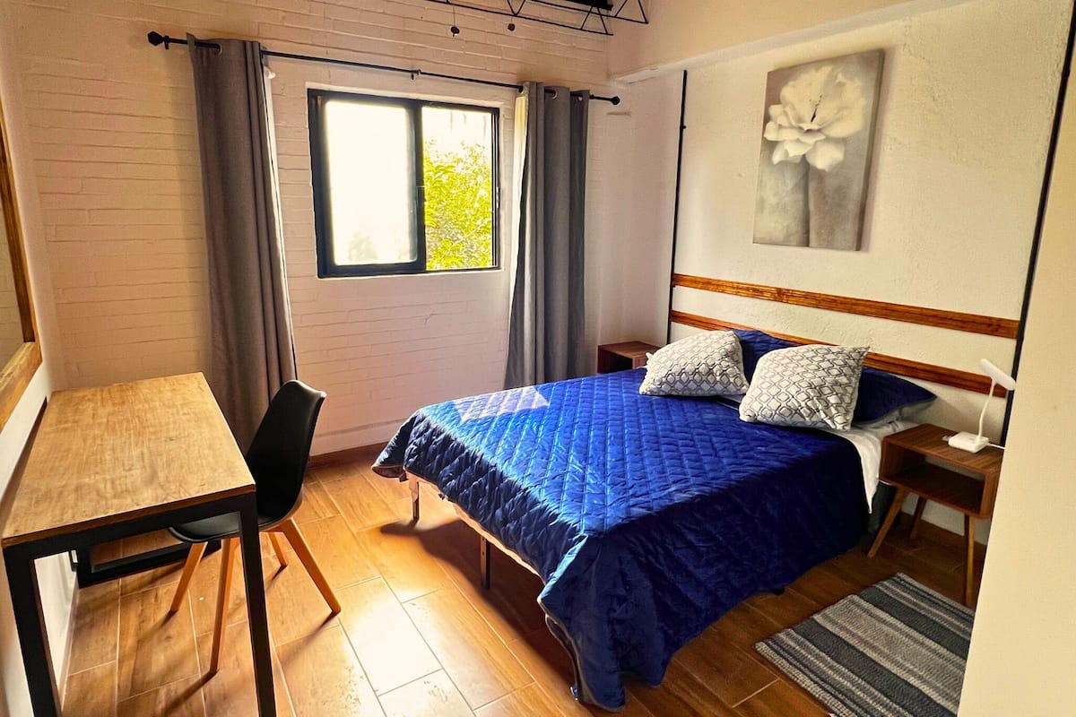 A bedroom with a bed, a desk, a chair, a window, and a painting on the wall.