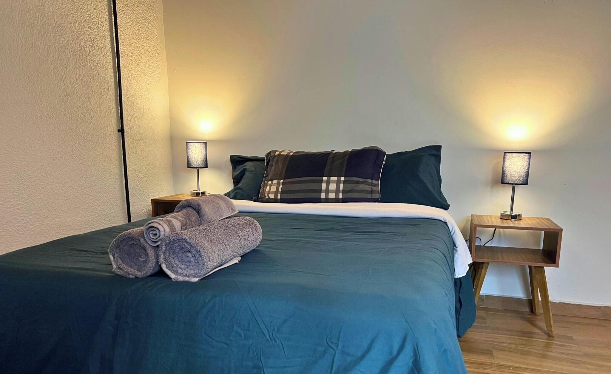 A neatly made bed with a teal bedspread, two pillows, and a rolled-up towel on the side. There are two bedside tables with lamps, one on each side of the bed.