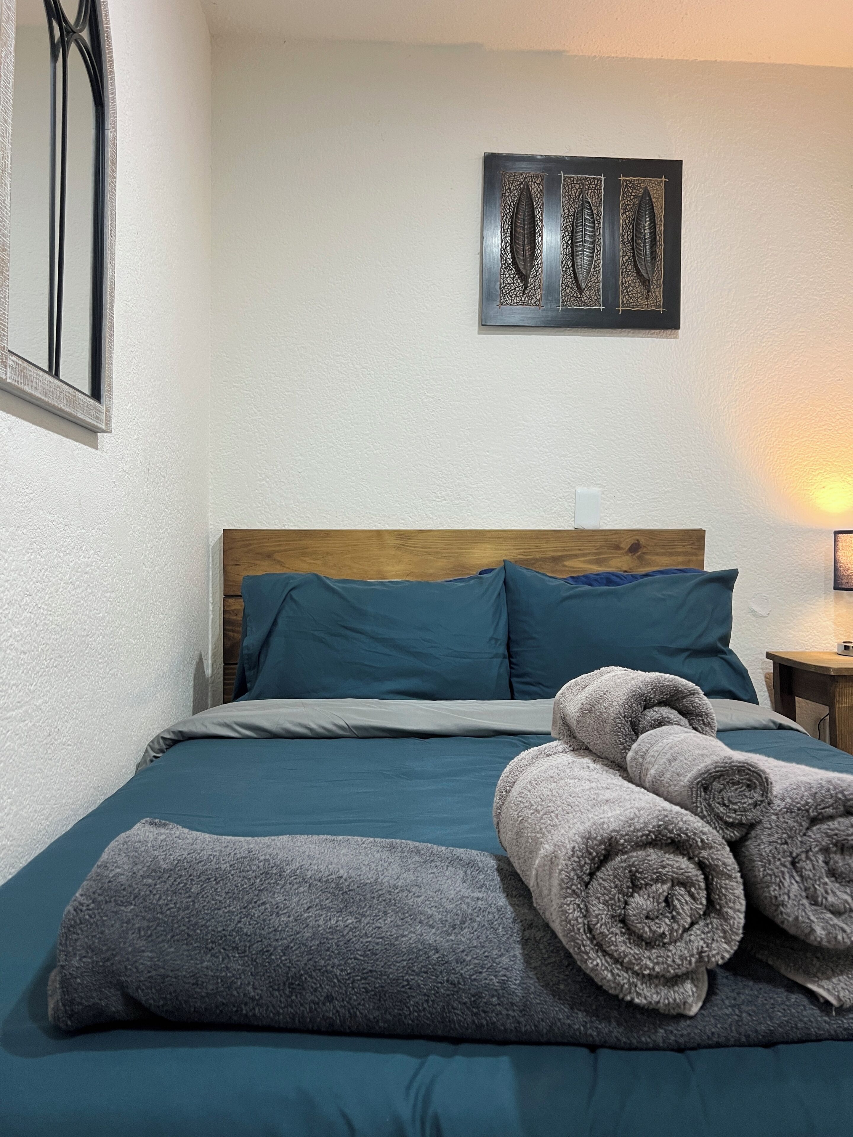 Een net opgemaakt bed met blauwgroene beddengoed, een houten hoofdbord en opgerolde handdoeken op het bed.