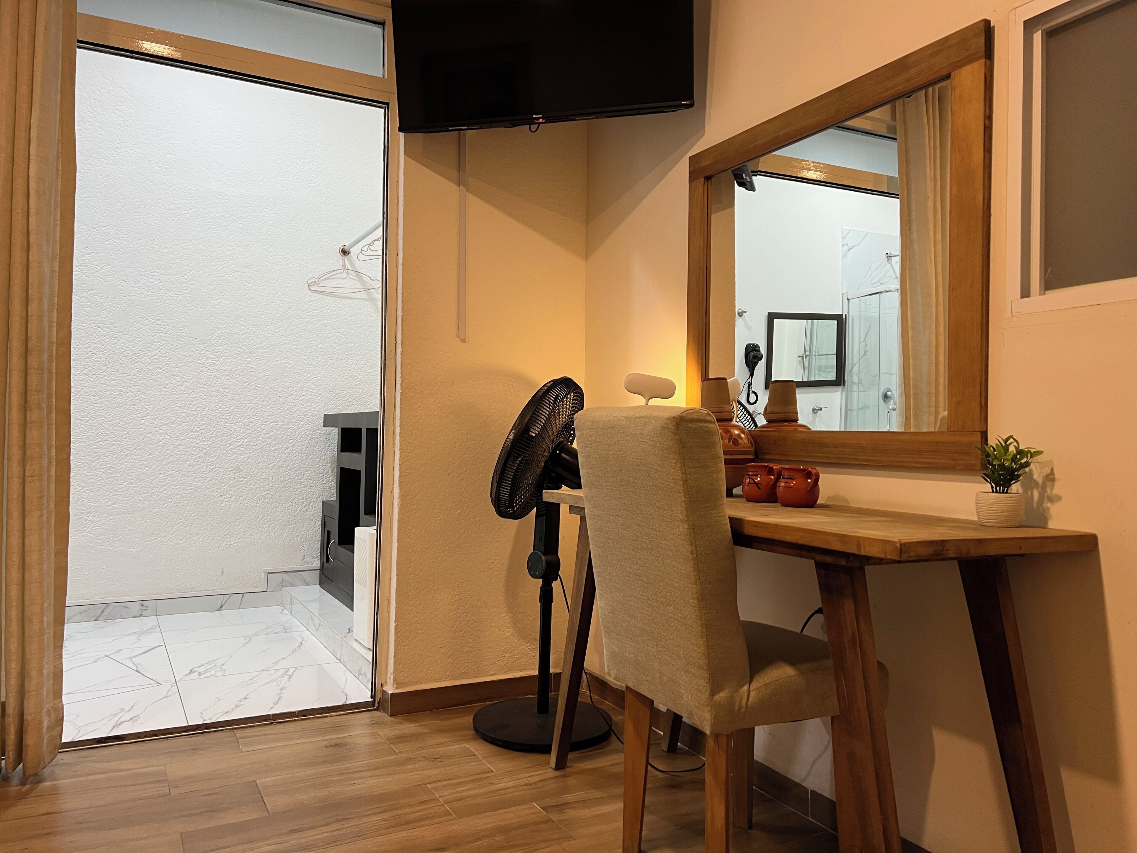 A room with a wooden desk, a chair, a standing fan, a television mounted on the wall, and a mirror.