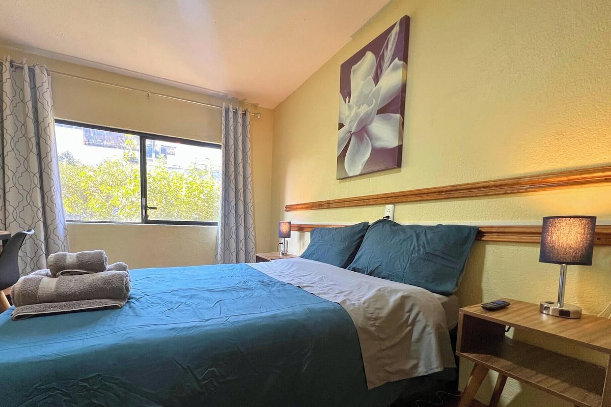 A hotel room with a bed, a desk, a chair, a lamp, and a painting on the wall.