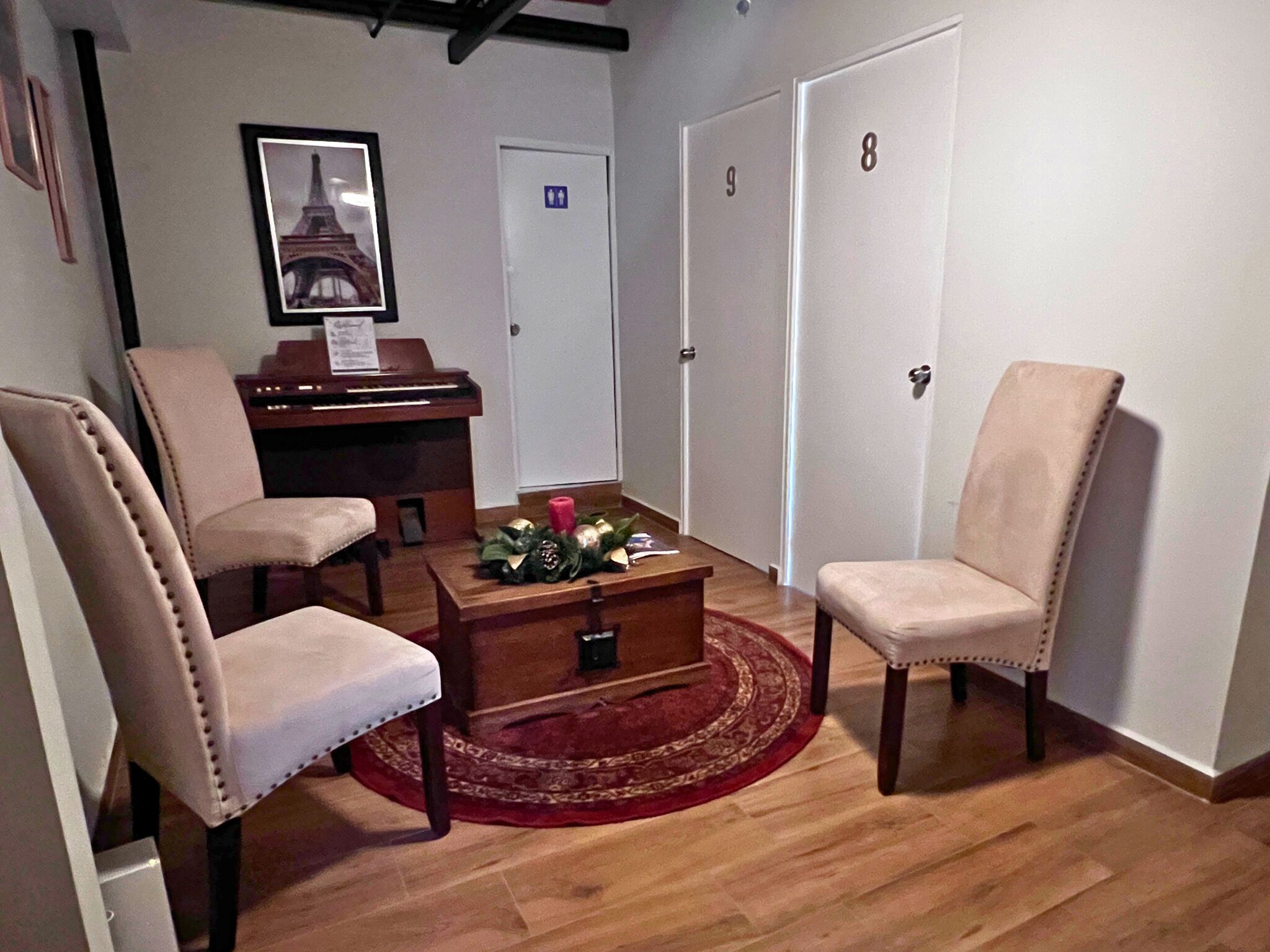 Une pièce avec un piano, deux fauteuils, une table basse surmontée d’un tapis rouge et une porte avec une pancarte.