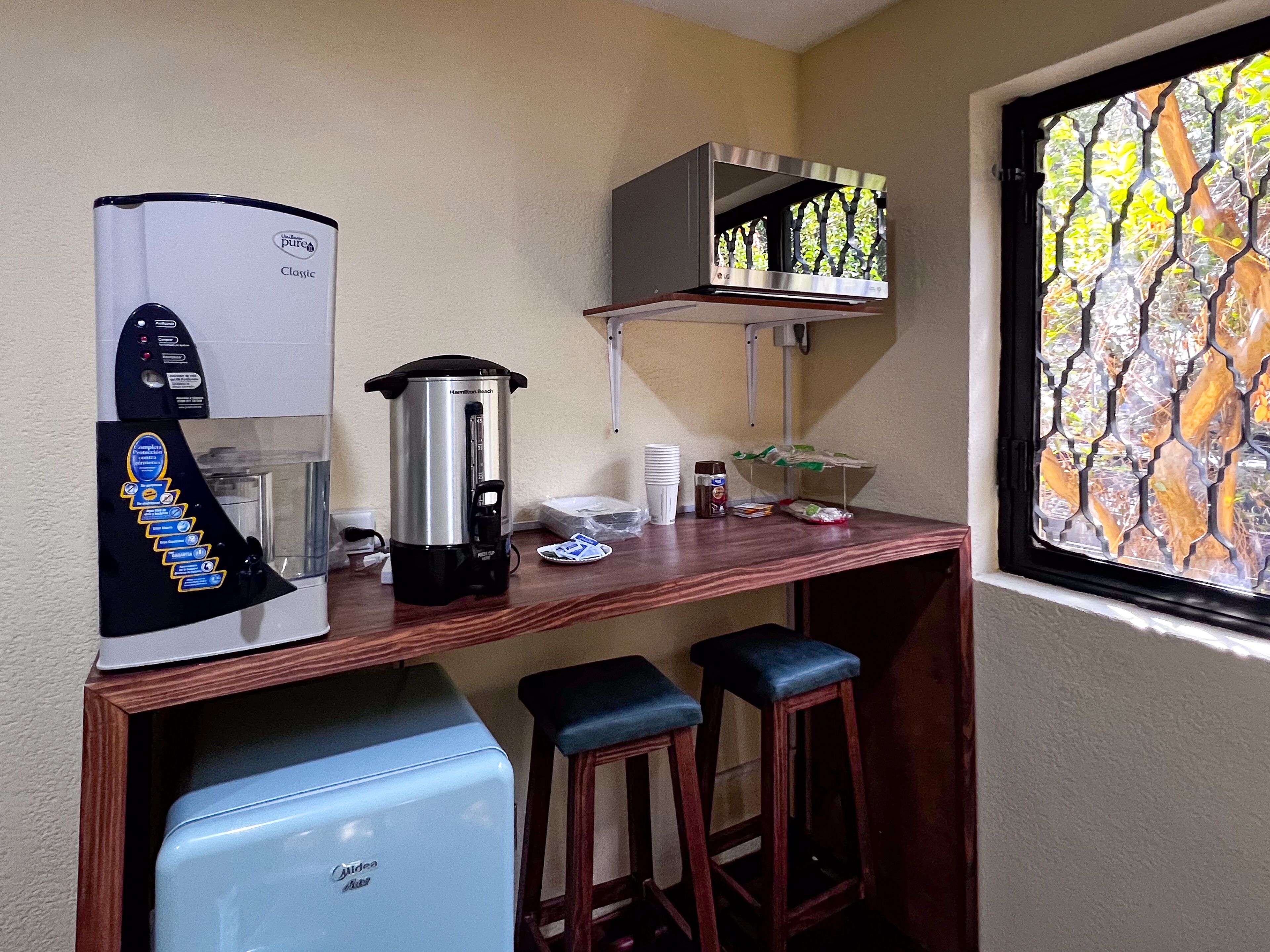 A kitchenette with a coffee maker, a kettle, and a mini fridge. There are two stools and a window with a view of trees.