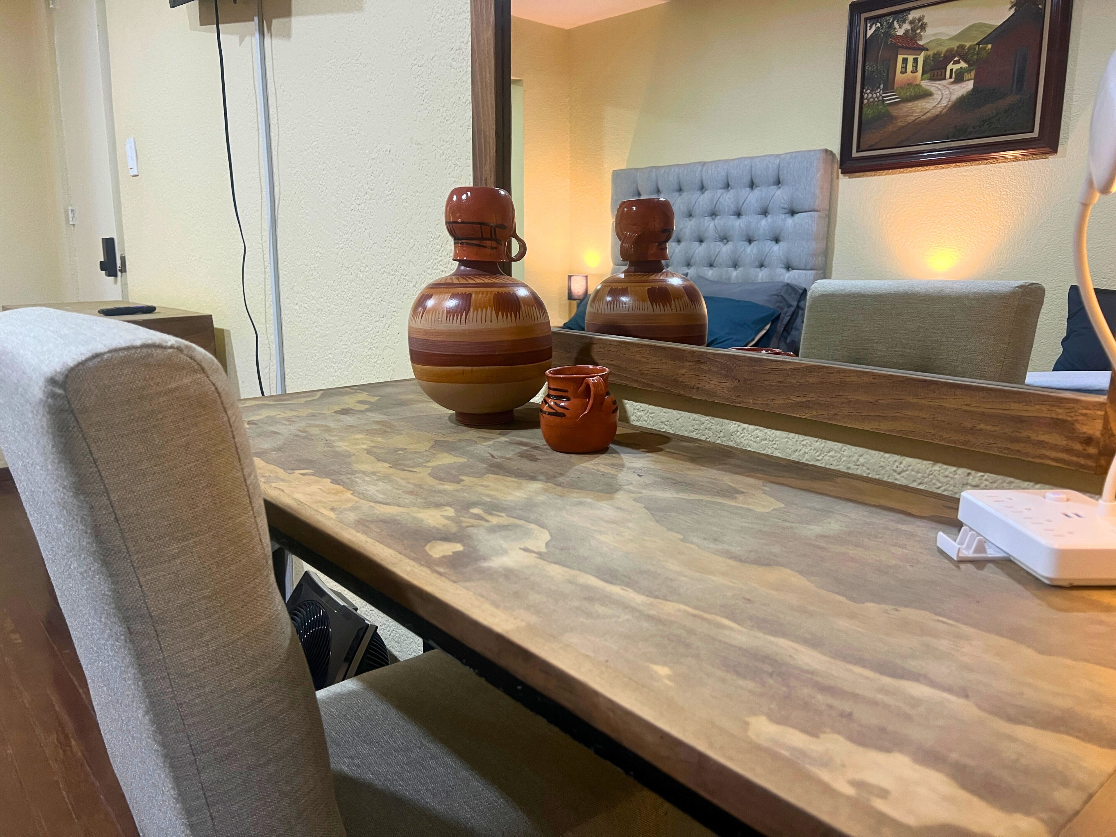 Une chambre d’hôtel avec une table en bois, deux vases en céramique, un miroir qui reflète la pièce, un lit avec une tête de lit capitonnée et un tableau accroché au mur.