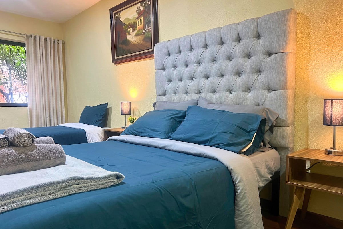 A hotel room with a large bed, two bedside tables, a nightstand, a lamp, a painting on the wall, and a window with curtains.