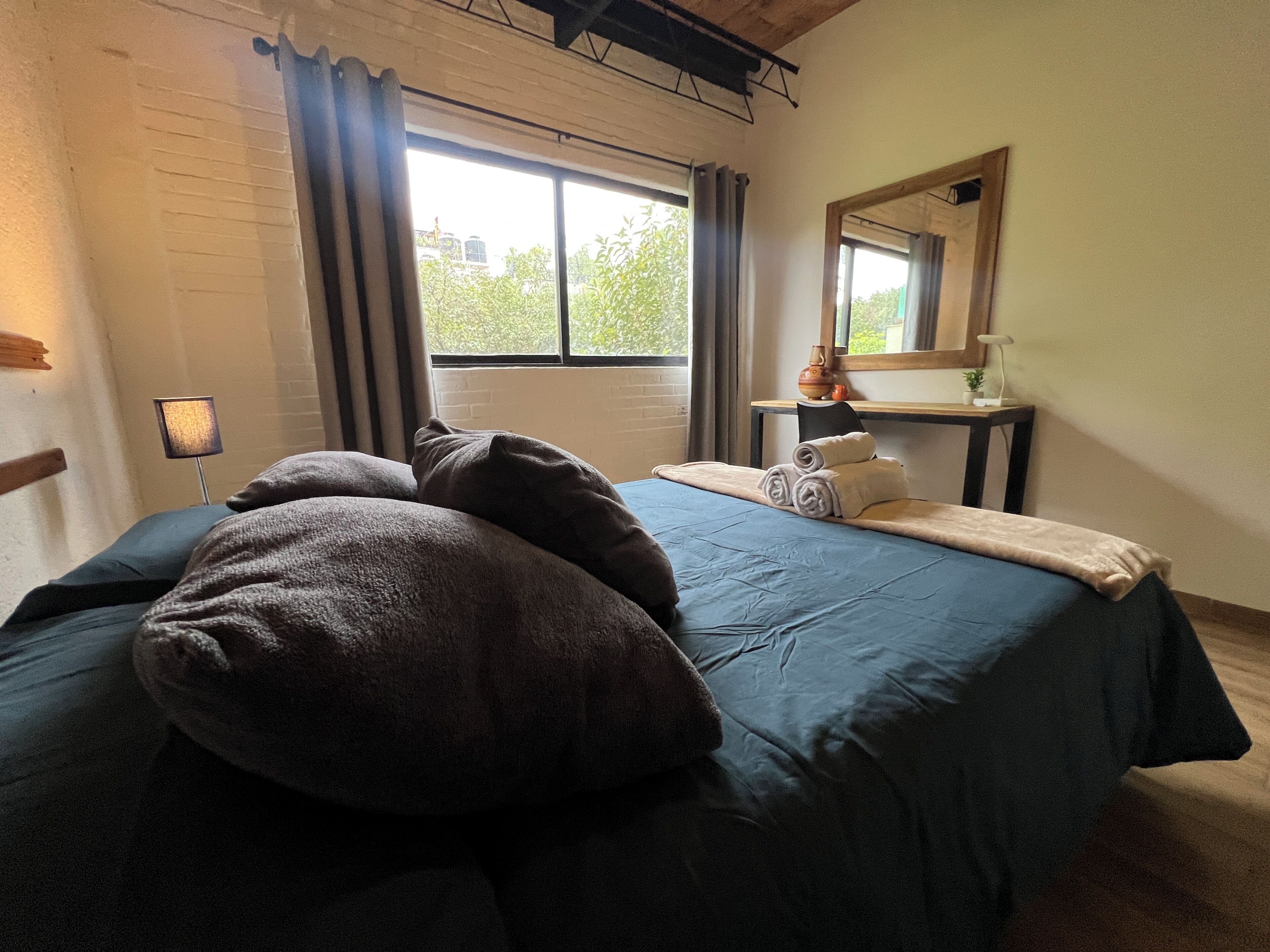 A bedroom with a bed, pillows, a nightstand with a lamp, a window with curtains, and a mirror.