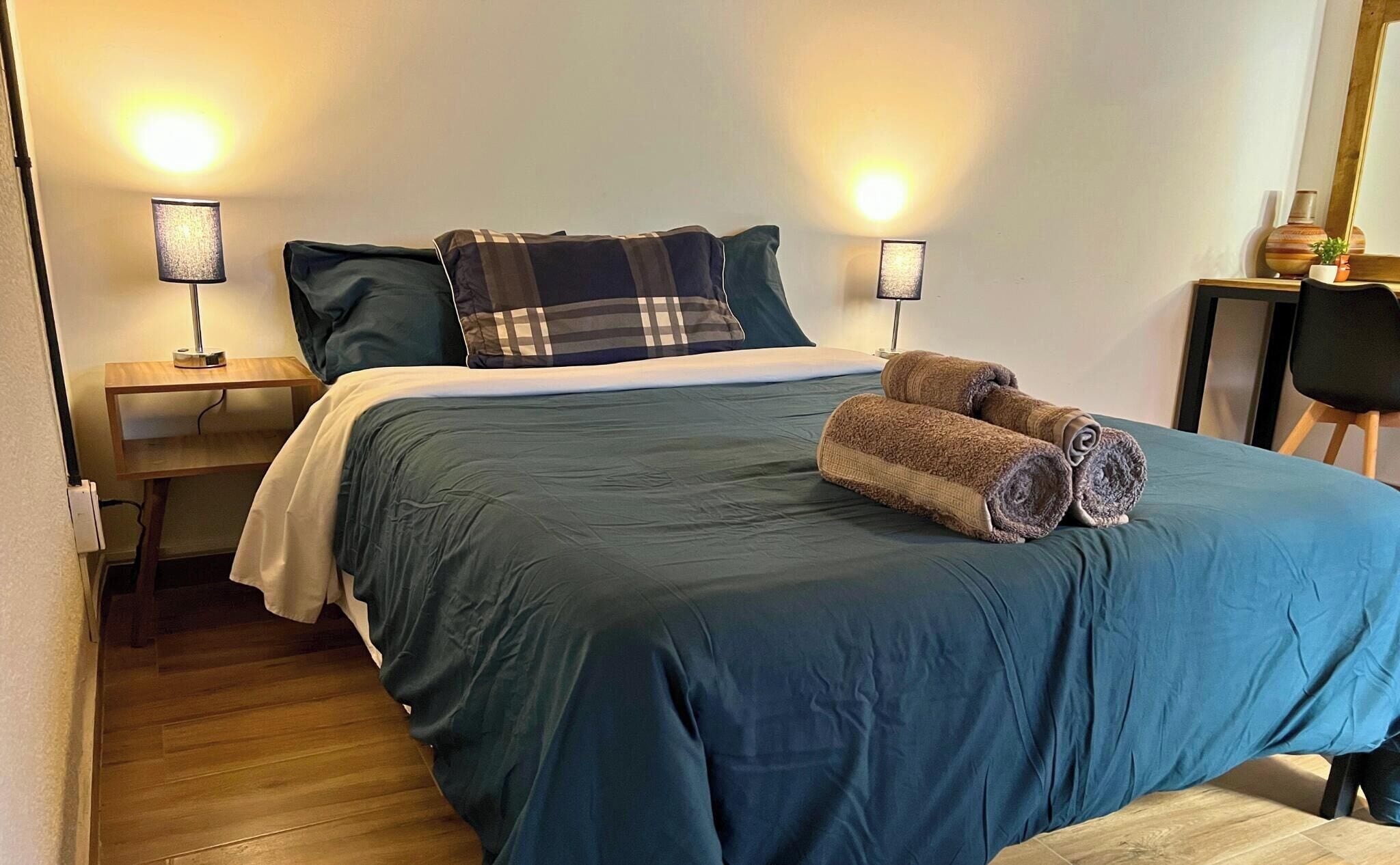A neatly made bed with a blue bedspread, two pillows, and rolled towels. There is a small wooden nightstand with a lamp, a chair, and a small table with a vase and plant.