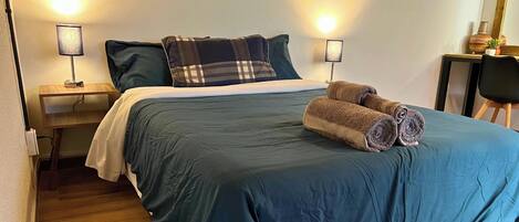 A neatly made bed with a blue bedspread, two pillows, and rolled towels. There is a small wooden nightstand with a lamp, a chair, and a small table with a vase and plant.