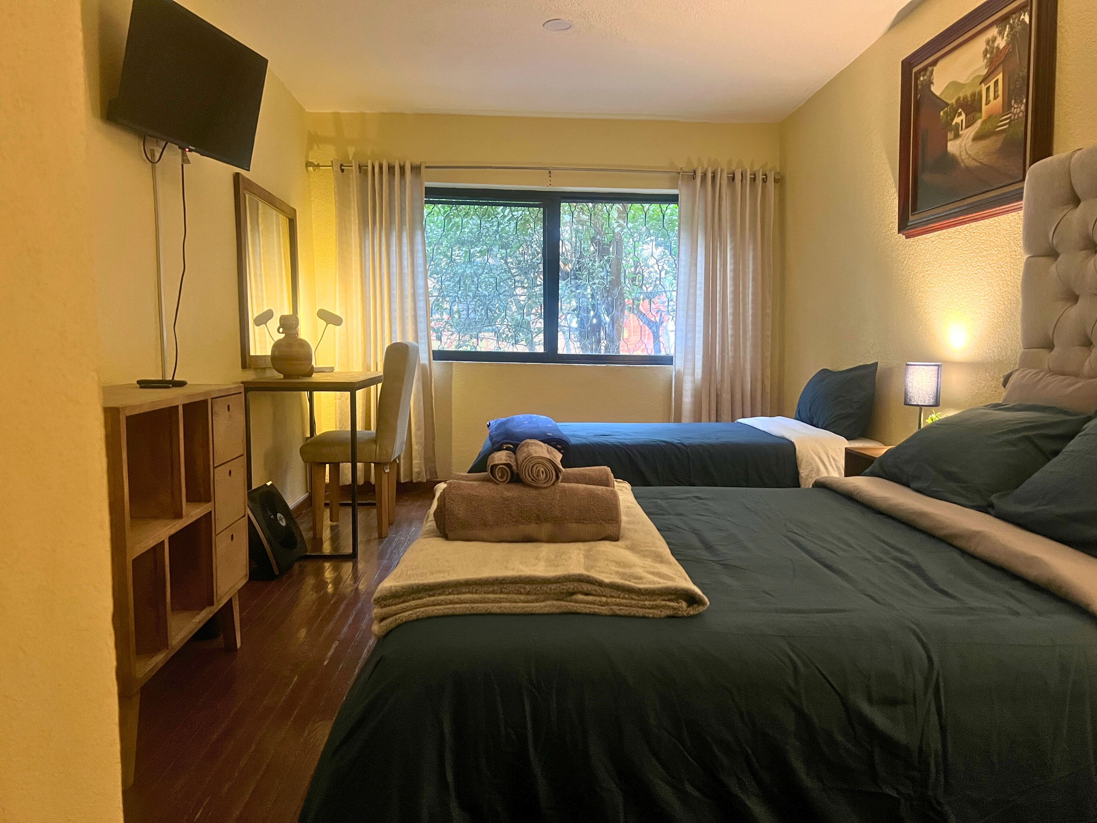 Habitación de hotel con dos camas, un escritorio con silla, televisión y ventana con cortinas.