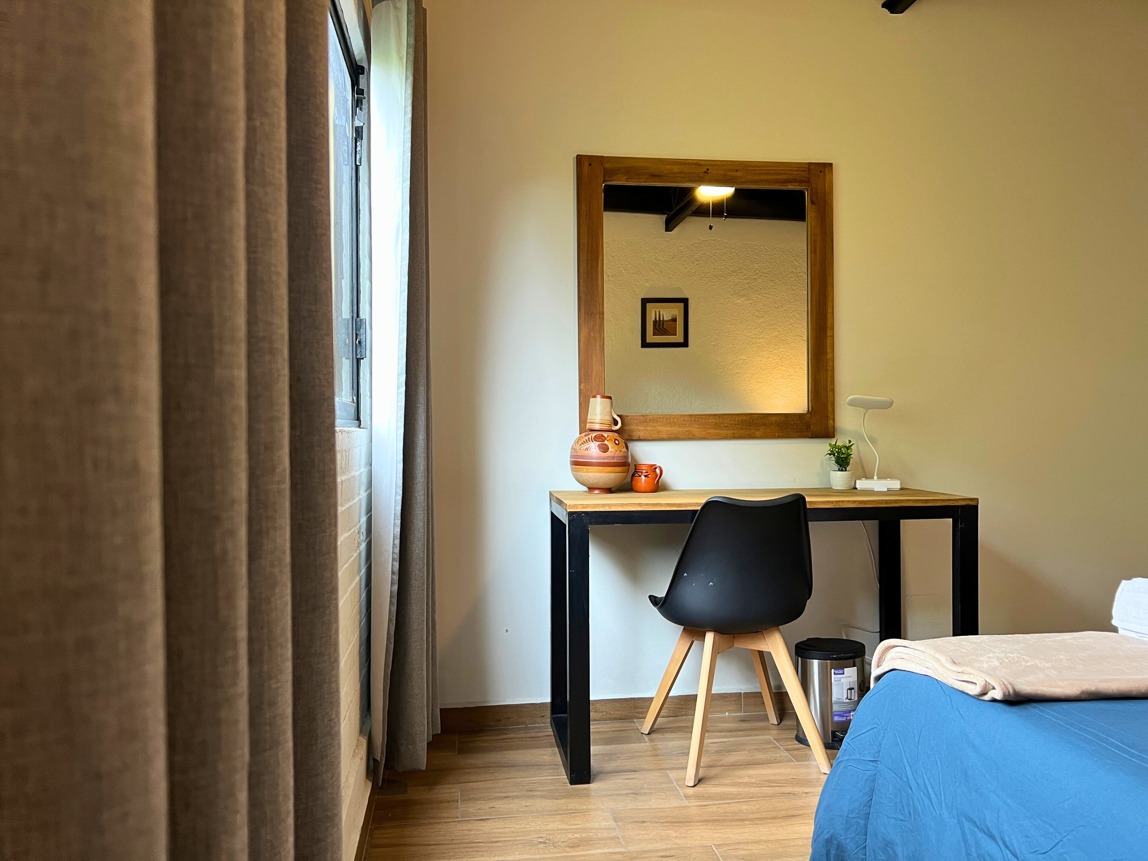 A room with a bed, a desk with a chair, a small potted plant, and a mirror on the wall.