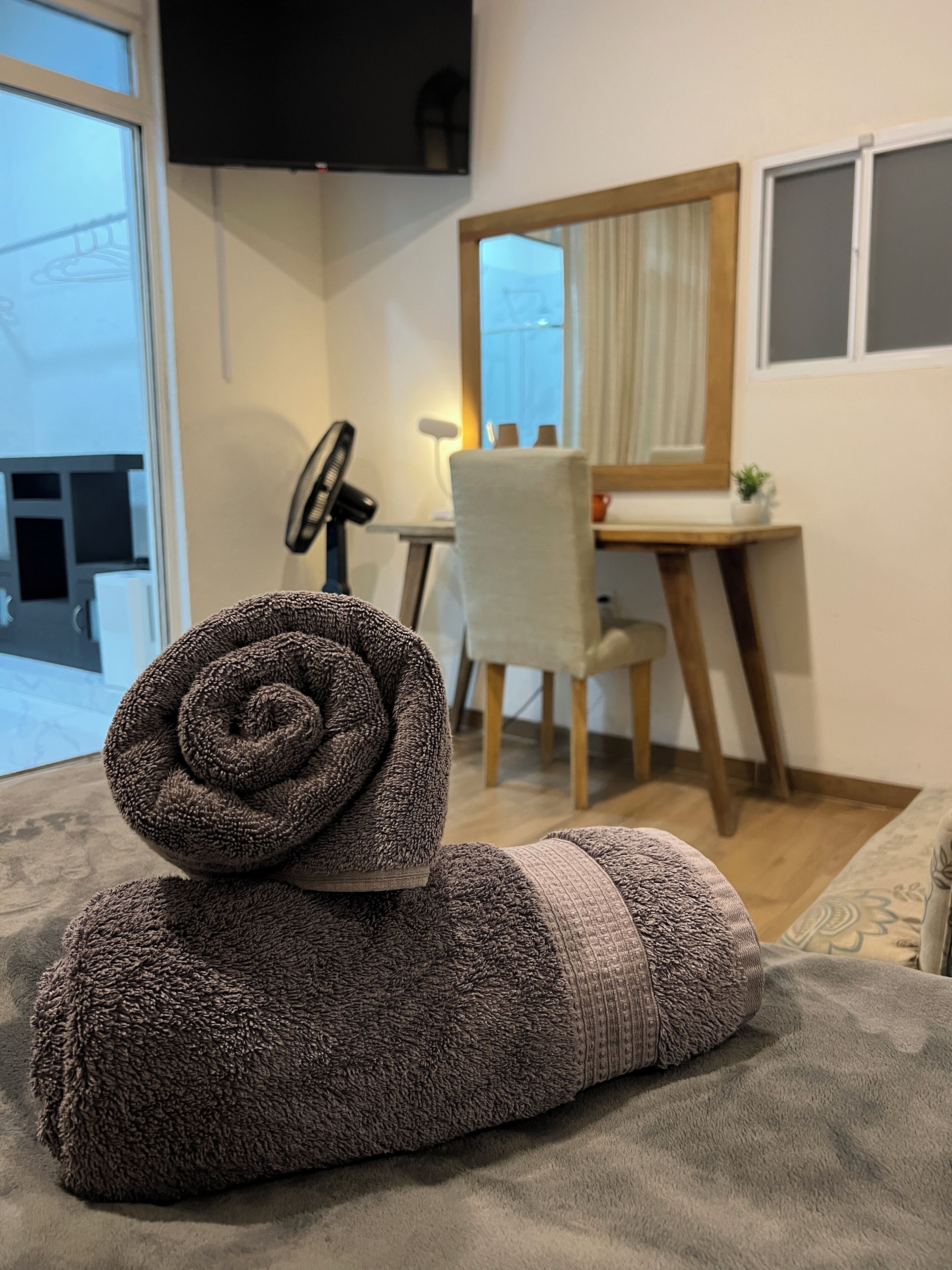 Ein ordentlich bezogenes Bett mit einem zusammengerollten Handtuch, ein Schreibtisch mit Stuhl, ein Spiegel und ein Fenster mit Vorhängen.