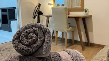 A neatly made bed with a rolled towel, a desk with a chair, a mirror, and a window with curtains.
