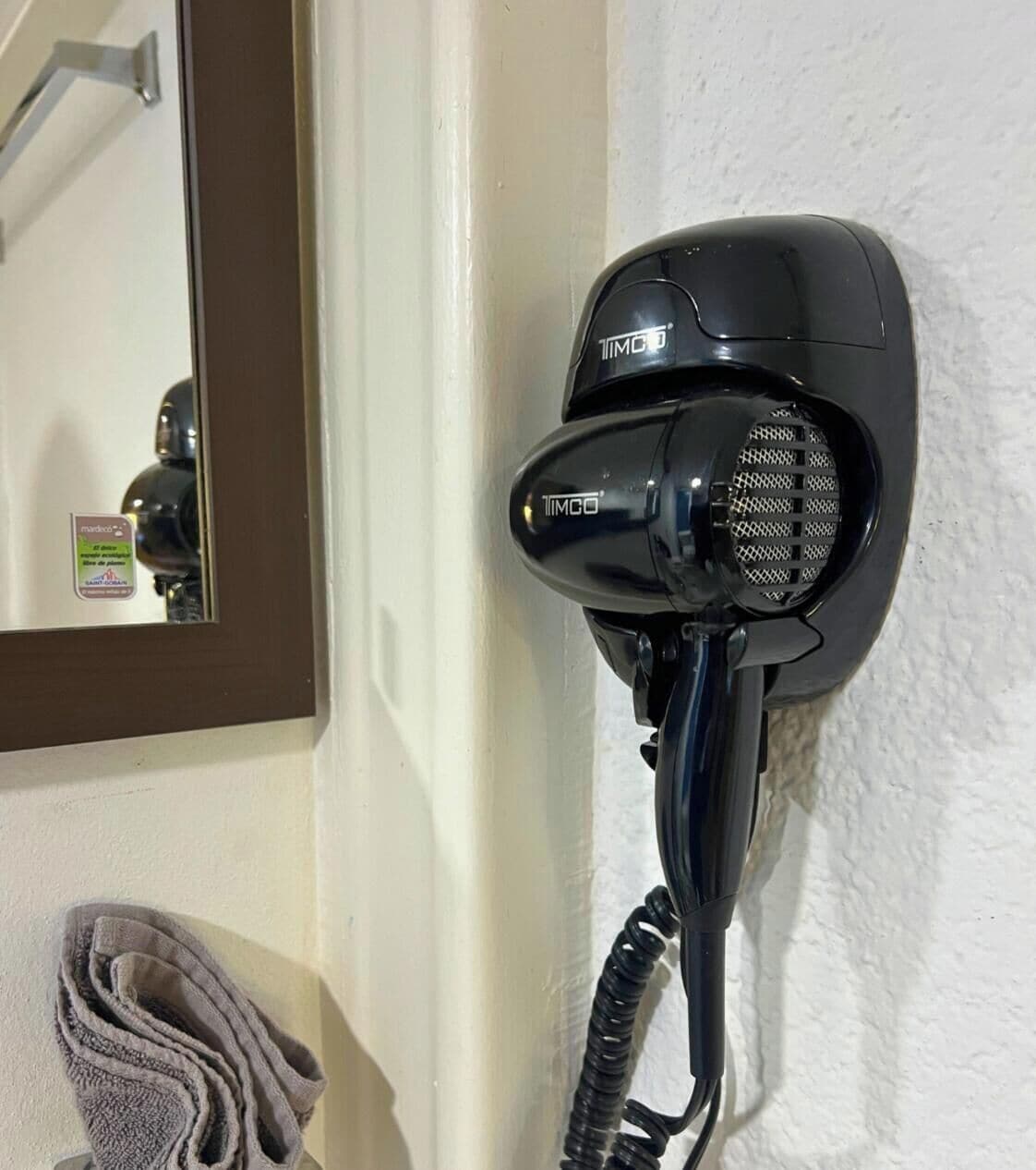 Ein schwarzer Föhn an der Wand befestigt, ein Handtuch an einem Haken und ein Spiegel, der einen Teil des Raumes reflektiert.