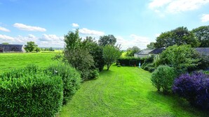 Un área verde bien cuidada con diversos árboles y arbustos, un cielo azul despejado y una vista distante de una casa.